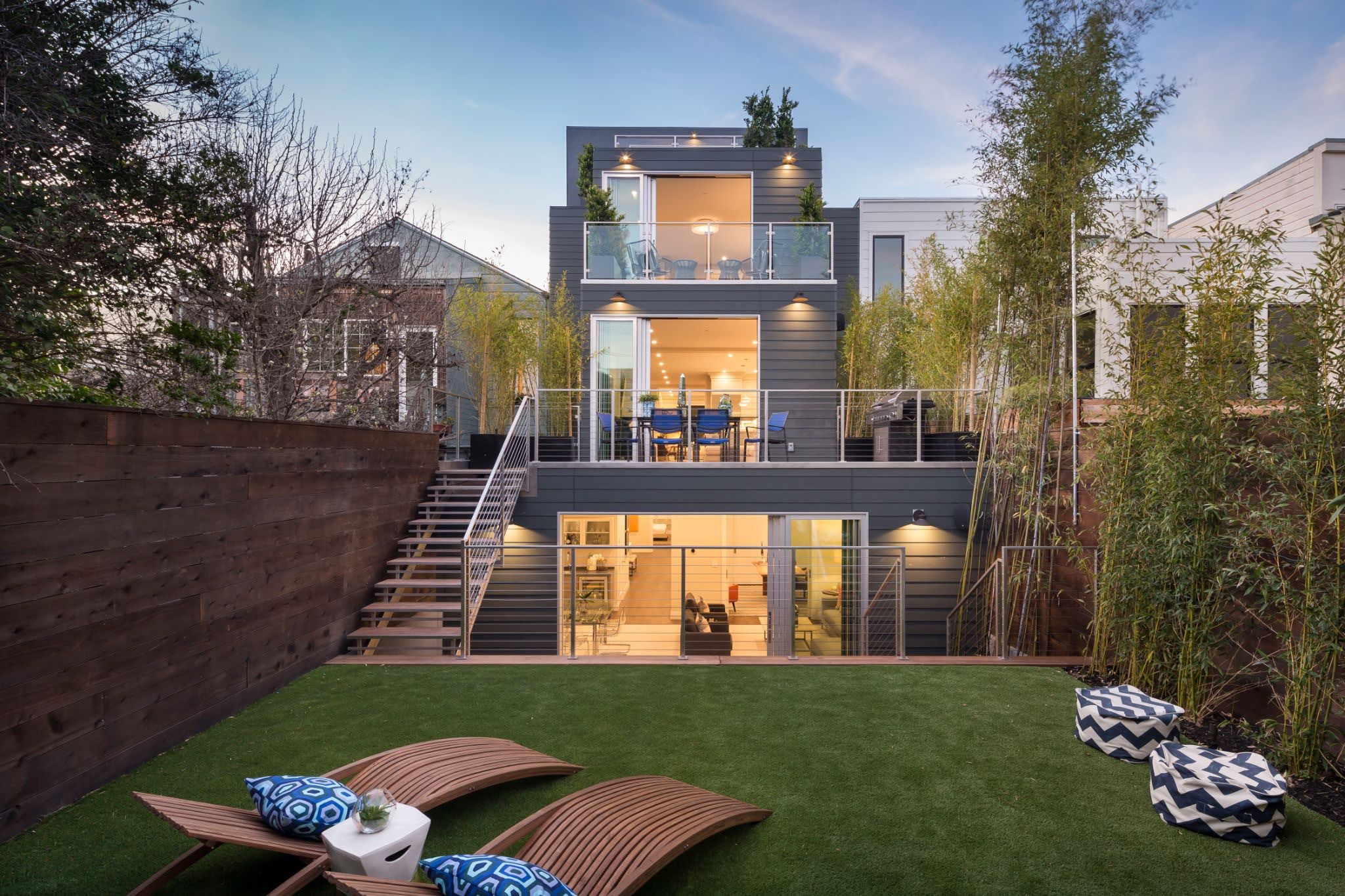 unnamed-18 Modern three-story house with large glass windows and terraces, viewed from a backyard with wooden deck chairs and pillows on artificial grass. Tall trees and fencing surround the space.