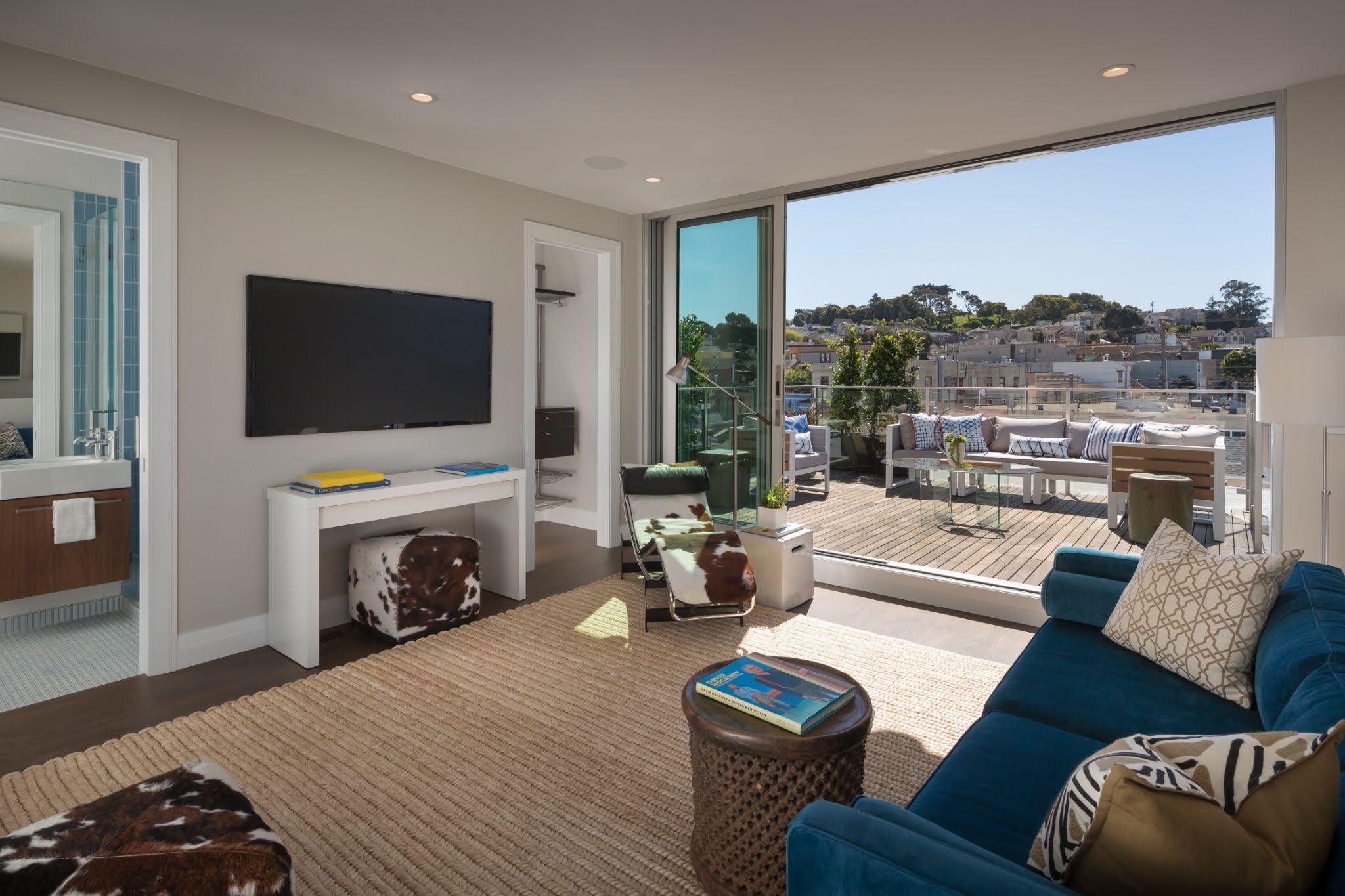 unnamed Modern living room featuring a wall-mounted TV, a blue couch, and a sliding glass door opening to a spacious balcony with outdoor seating. A bathroom is visible to the left through an open door.
